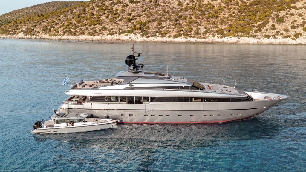Sleek silver NAIA yacht anchored in calm waters near a rocky Greek coastline with a tender alongside.